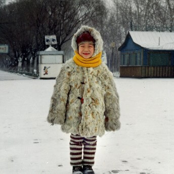 Still from Xmas Without China. A young child stands outside in the snow. They wear snow boots, striped pants, a fur coat, and a yellow scarf. A blue house and a white stand are situated behind them.