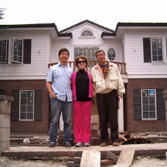 Still from Xmas Without China. From left to right, a young man wears a blue and white blouse with jeans, a woman wears a pink jumpsuit with a black top, and an older man wears black pants and a white jacket. They stand in front of a white and brick house with construction materials surrounding them.