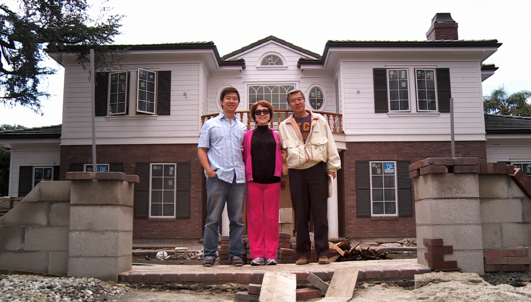 Still from Xmas Without China. From left to right, a young man wears a blue and white blouse with jeans, a woman wears a pink jumpsuit with a black top, and an older man wears black pants and a white jacket. They stand in front of a white and brick house with construction materials surrounding them.