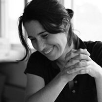 Holen Sabrina Kahn laughing and looking down while holding both of her hands together in front of the camera. Her hair is pulled up on the back of her hair, and she wears a polo shirt. Portrait in black and white.