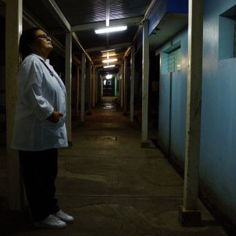 Still from A Quiet Inquisition. Profile full shot of a doctor wearing a doctor robe, and an stethoscope. She is stanfing against a column during night time. The shot is taken from a vanishing point of view.