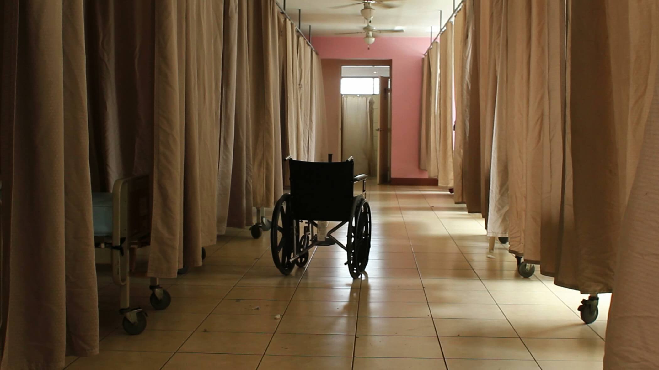 Still of A Quiet Inquisition. Shot of a wheelchair in a hospital room with curtains between each bed.