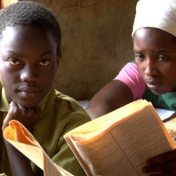 Still from A Small Act. A shot of two children, one of them looks at the camera, while the other one, a girl, reads something from a book. It is daytime.