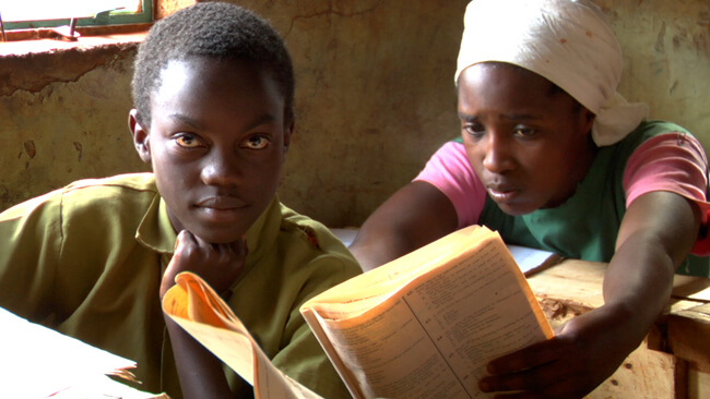 Still from A Small Act. A shot of two children, one of them looks at the camera, while the other one, a girl, reads something from a book. It is daytime.