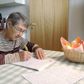 Still from A Small Act. A shot of an elderly woman sitting and writing on paper on a table in a kitchen, there are more papers with her handwriting, and there is also a basket on the table. The woman has short gray hair and glasses. It is daytime.