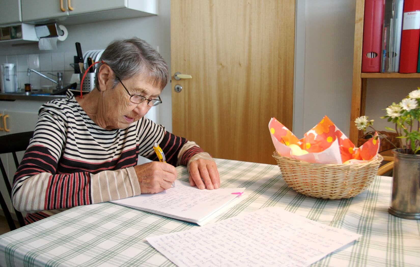 Still from A Small Act. A shot of an elderly woman sitting and writing on paper on a table in a kitchen, there are more papers with her handwriting, and there is also a basket on the table. The woman has short gray hair and glasses. It is daytime.