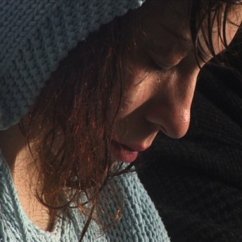 Still from About Face: The Story of Gwendellin Bradshaw. Profile close-up to Gwendellin Bradshaw, she wears a knitted baby blue hoodie and looks down.