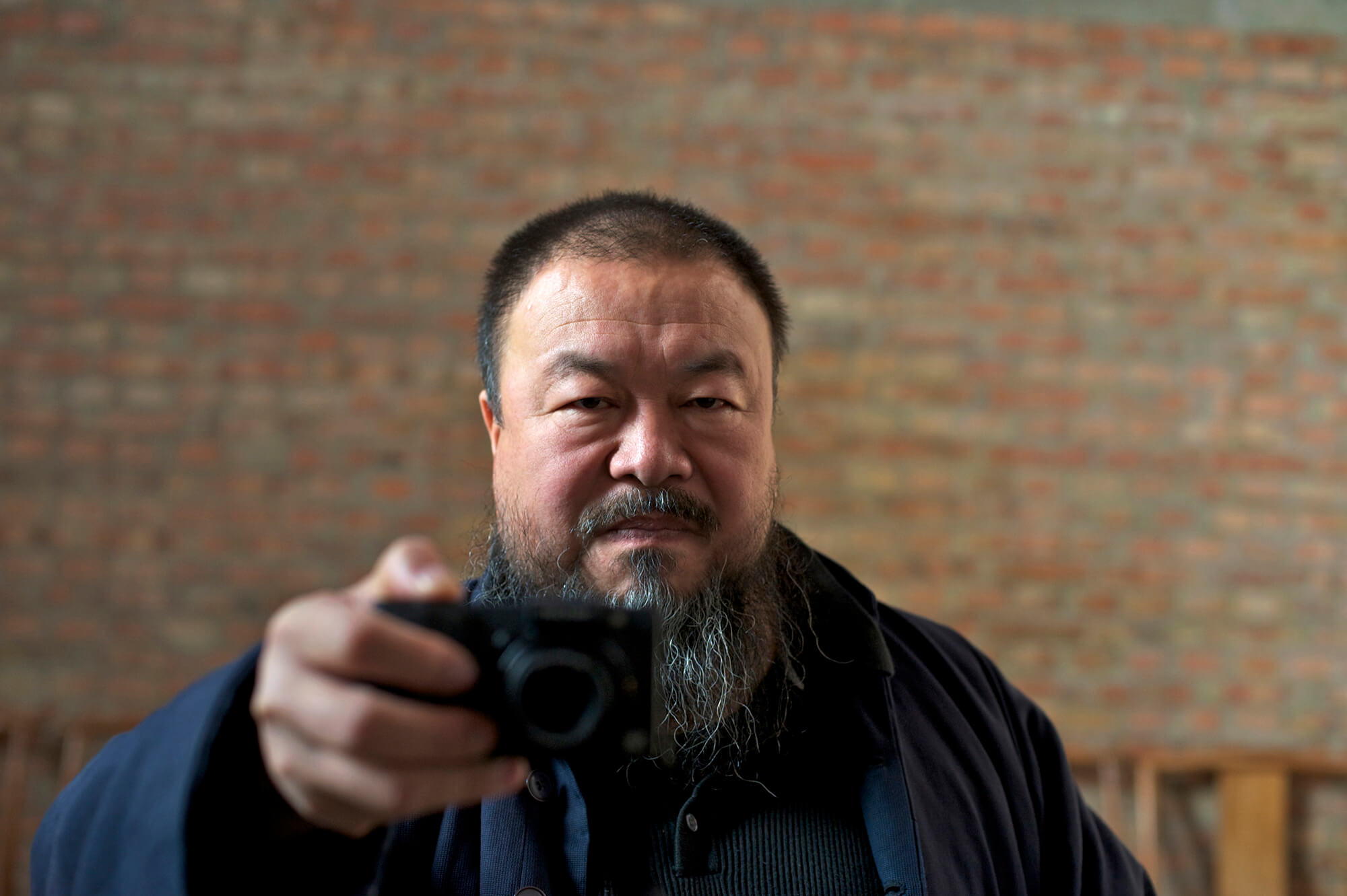 Selfie of Ai Wei Wei. He is aiming with a camera in front of a mirror.