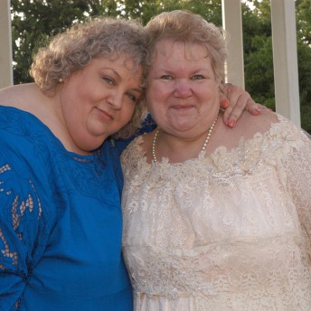 Still from All of Me. Two women pose in front of the camera, the woman on the left has a blue dress and hugs the woman to her right. The woman on the right is wearing a pearl-white dress, and smiles at the camera.