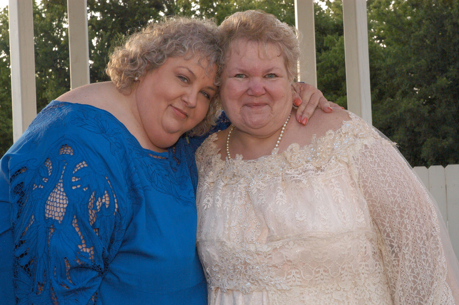 Still from All of Me. Two women pose in front of the camera, the woman on the left has a blue dress and hugs the woman to her right. The woman on the right is wearing a pearl-white dress, and smiles at the camera.