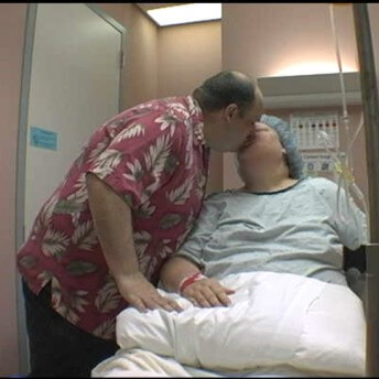 Still from All of Me. In a hospital room, a couple is kissing; a woman is in hospital clothing and sitting on a bed; a man is standing and leaning in to kiss her.