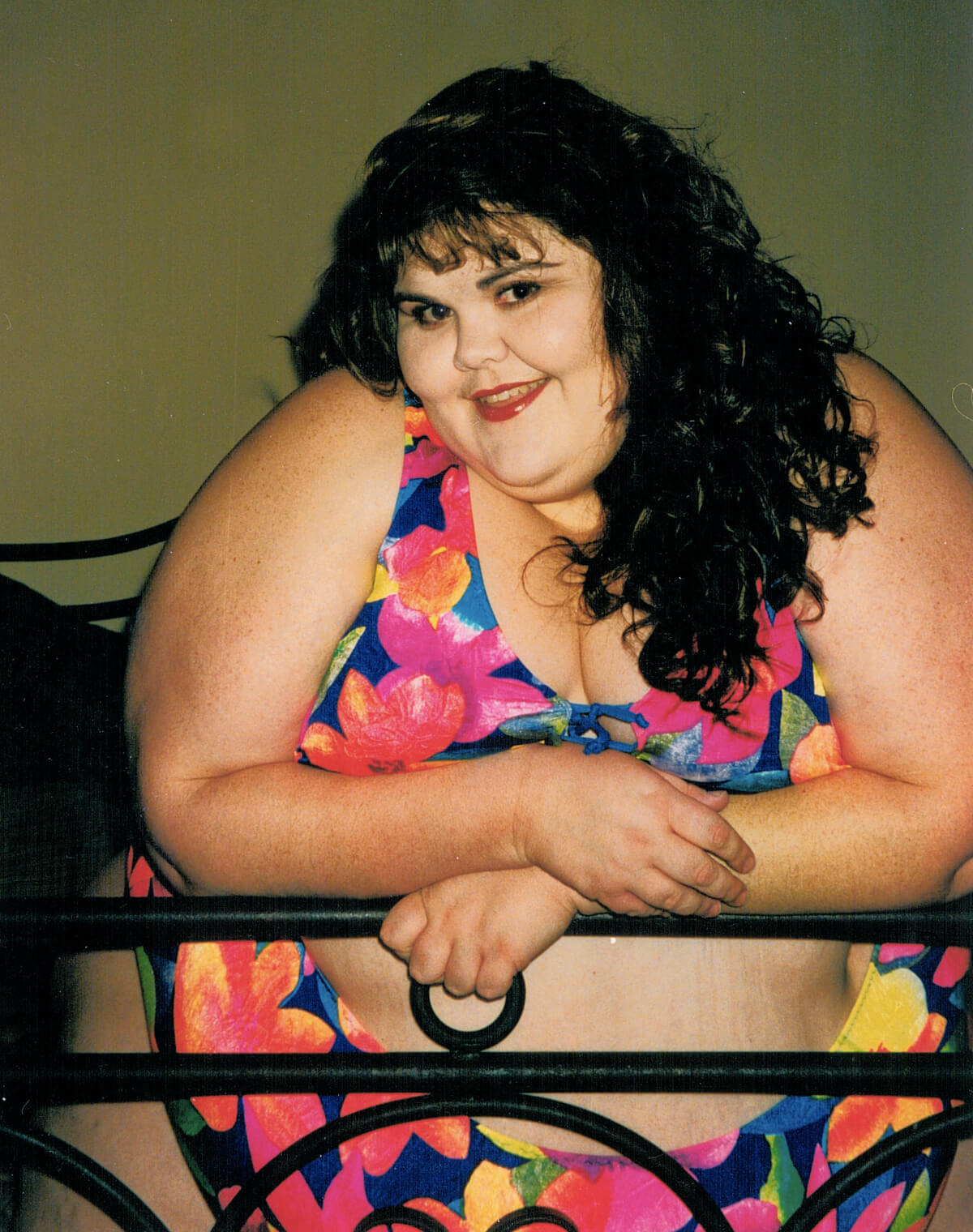 Still from All of Me. A woman wearing a bikini poses at the camera, she is leaning on a bed frame, and smiling.