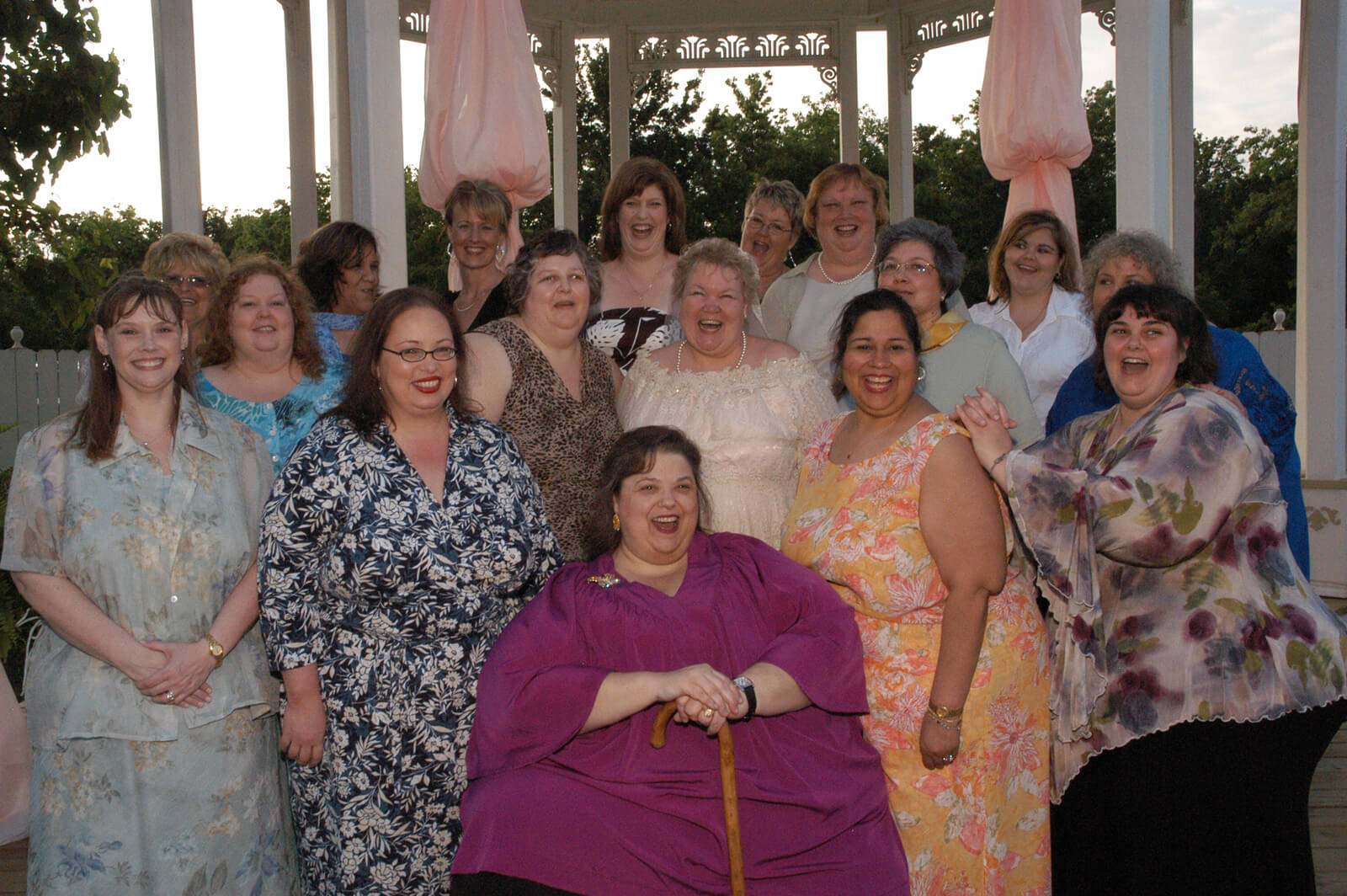 Still from All of Me. Group photo of sixteen women with dresses smiling at the camera.