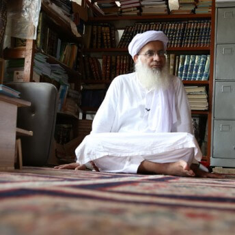 Still from Among The Believers. A man with a white beard and wearing a turbin is sitting with his legs crossed.