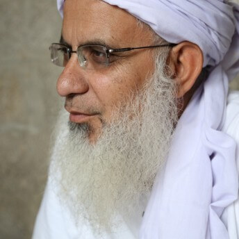 Still from Among The Believers. Profile shot of a man with a long gray beard, wearing eyeglasses, a white turban, and a white shirt.
