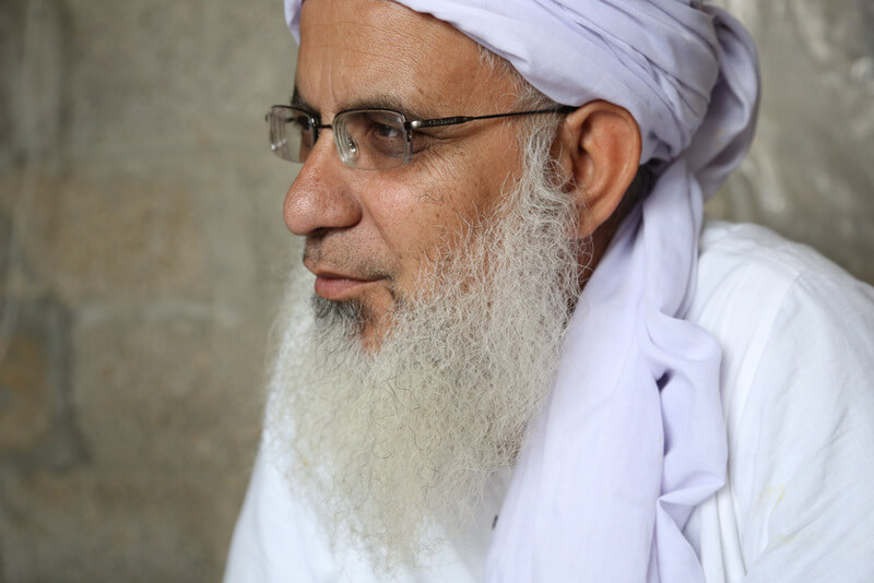 Still from Among The Believers. Profile shot of a man with a long gray beard, wearing eyeglasses, a white turban, and a white shirt.
