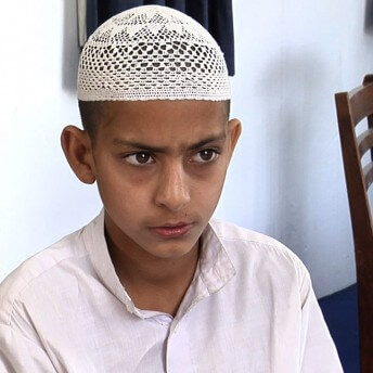 Still from Among The Believers. Medium shot of a child sitting on a chair, he looks away from the camera and wears a knitted-skull cap.