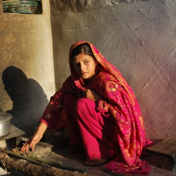 Still from Among The Believers. A woman with dark hair, kneeling on the floor to cook, in front of her there is a ground-level fire pit and a cooking pot. She wears a traditional Pakistani red dress.