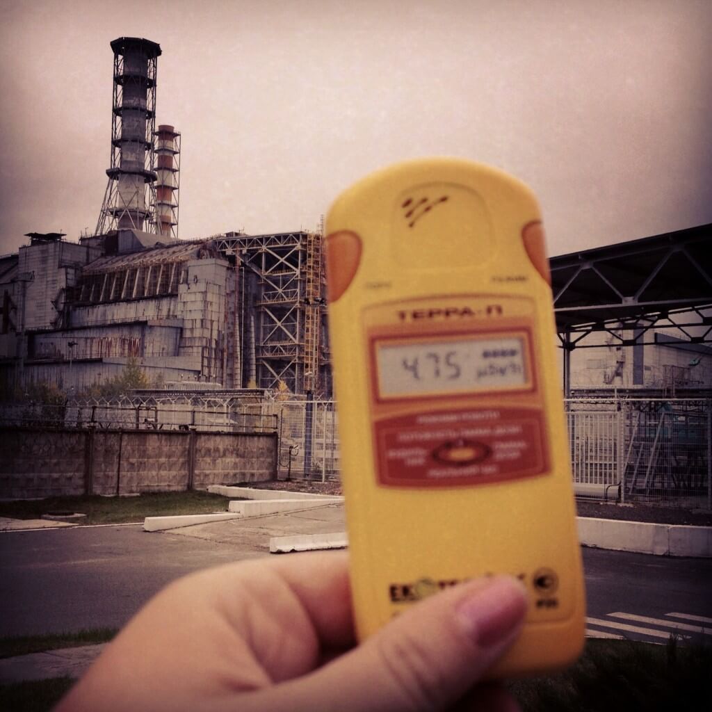 A photo of someone holding a yellow radiation monitor in front of an industrial plant. The monitor on the screen reads "475".