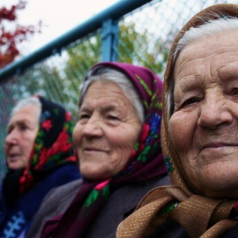 A close up shot of three elederly woman sitting next to each other. The one in the foreground looks directly into the camera, while the other two look off into the distance. They are all wearing headscarves.