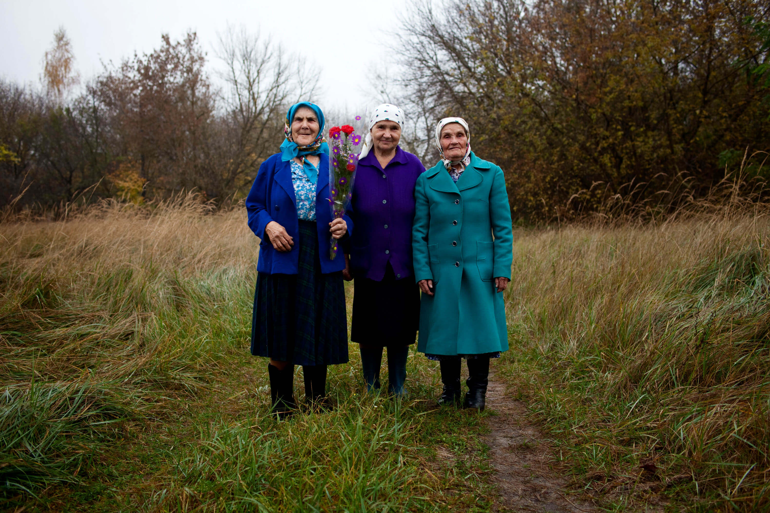Three women stand next to each other in the middle of a path that cuts through a grassy field. They are all looking directly at the camera. Each of them wears mid-calf dresses and skirts with jackets and big boots, all in various hues of black, blue, purple, and white. Each of them wears a headscarf.