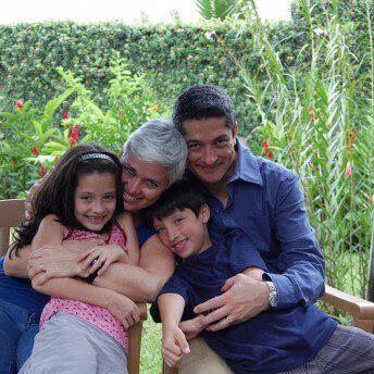 Still from Beautiful Sin. A family of a woman, a man, and two children smile at the camera. Parents are hugging the children. They all are in a garden.