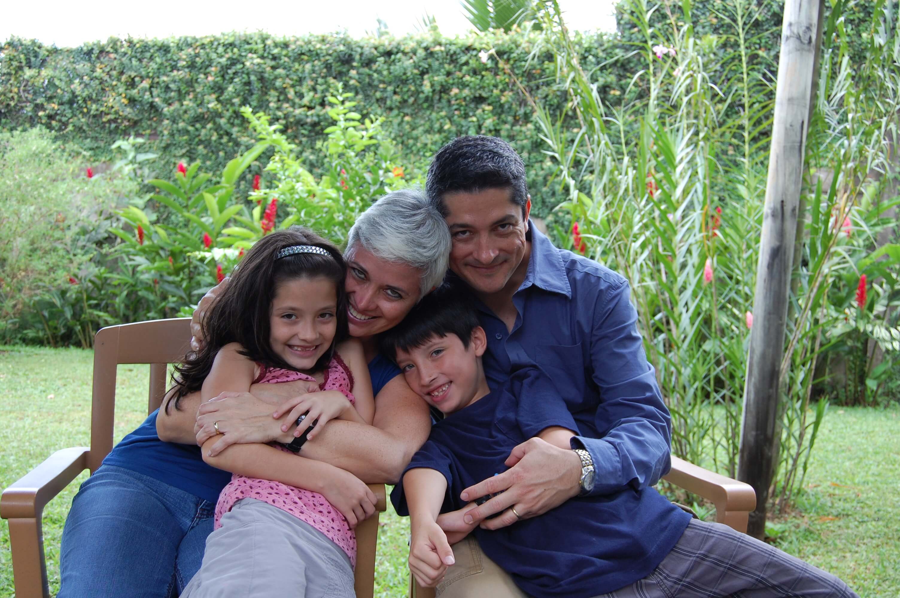 Still from Beautiful Sin. A family of a woman, a man, and two children smile at the camera. Parents are hugging the children. They all are in a garden.