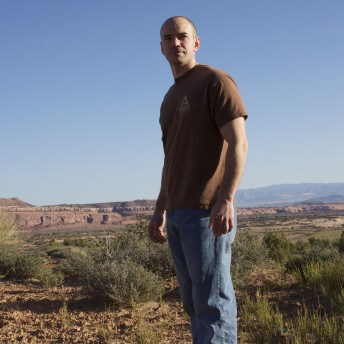A man wearing a t-shirt and jeans is standing in a desert landscape, behind there is a blue sky and desert vegetation.