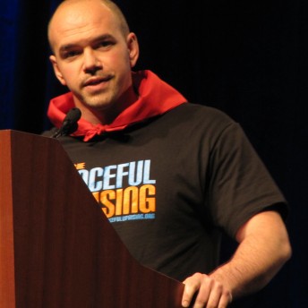 A man wearing a t-shirt is speaking behind a podium with a microphone.