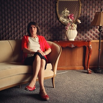 Still from The Pearl. A woman is sitting on a sofa, behind her there is a table with a vase with flowers, a mirror, and a lamp. The wall in the background has wallpaper.