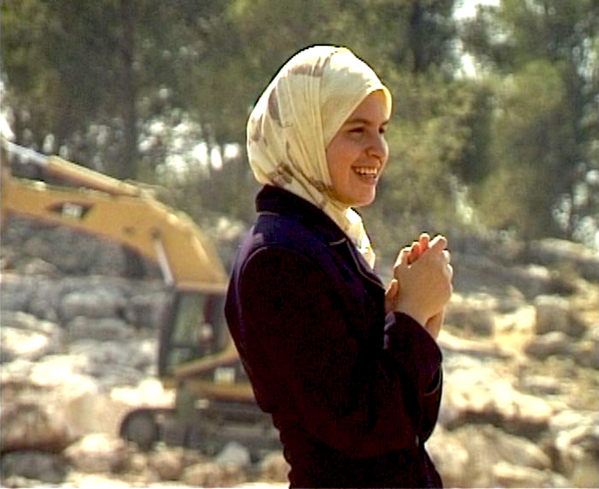 A woman is standing on a construction site, she is wearing a yellow hijad and holding her hands together in front of her chest.