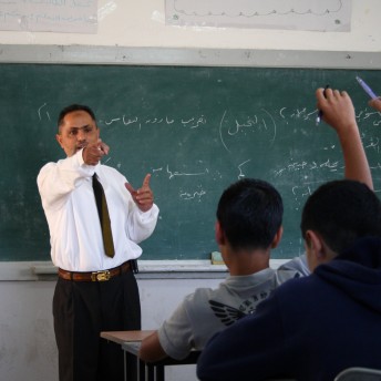 A classroom with a teacher and students. The professor is in front of the chalkboard and students are participating raising a their hands.