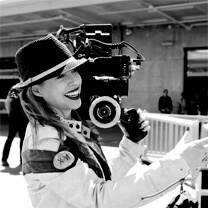Tiffany Shlain smiling and looking directly at the camera. She wears a jacket and a hat and has a camera on her shoulder. Black and white portrait.