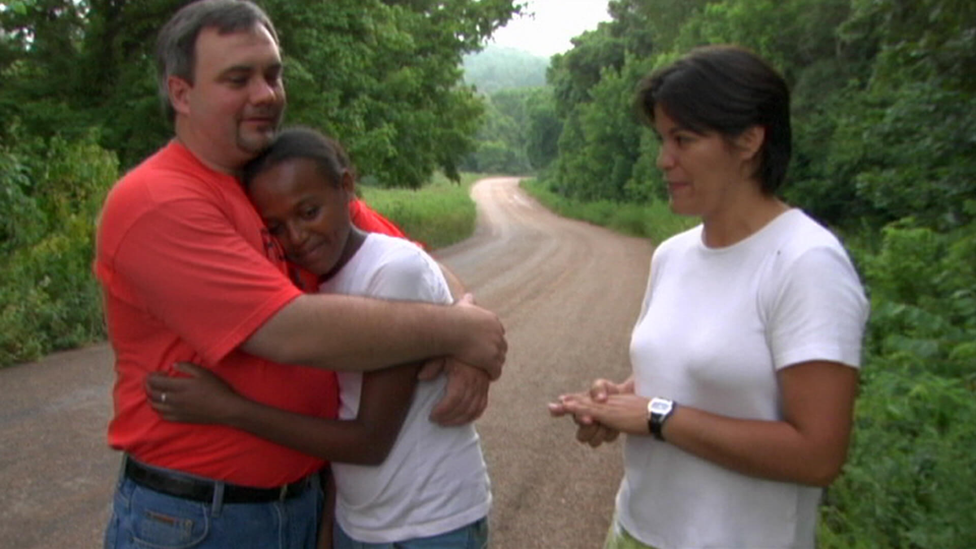 Still from Girl Adopted. A man and an adolescent girl are hugging, and a woman is watching them. Behind them is seen a winding dirt road, flanked by greenery on both sides.