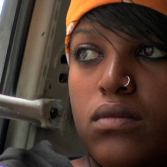 Still from Girl Adopted. Close up of young woman starting out of a window. She is wearing a nose ring and orange bandana.