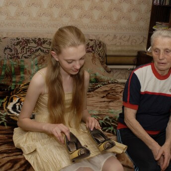 Still from Girl Model. A young girl with long blonde hair and wearing a light yellow dress, is looking at a pair of yellow and black heels that she is holding. She is sitting on a bed next to an elderly woman who is also looking at the shoes.