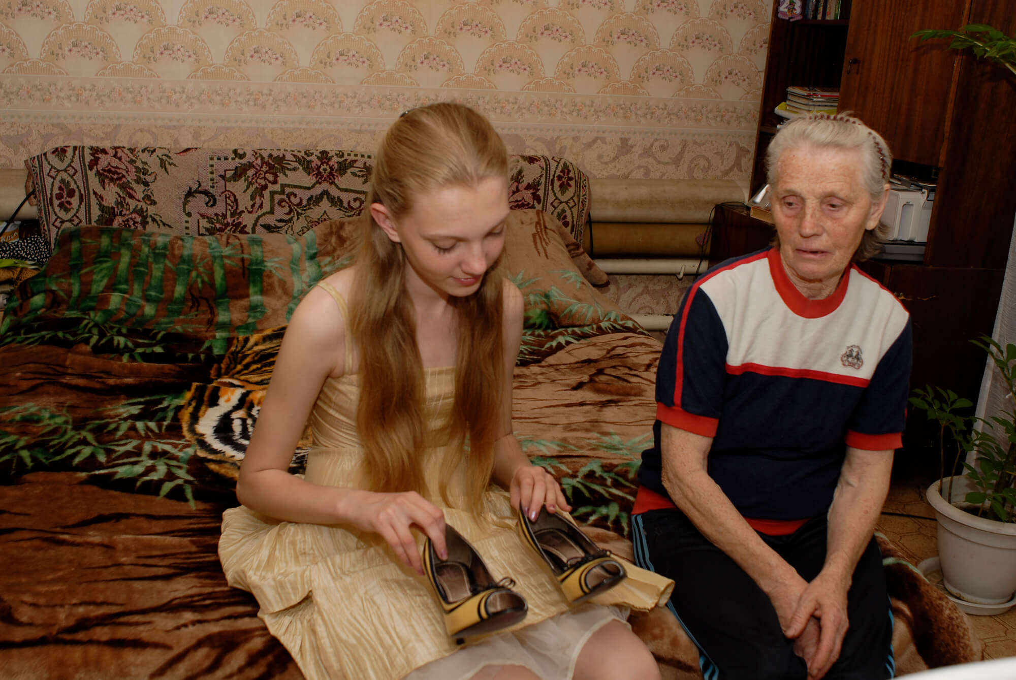 Still from Girl Model. A young girl with long blonde hair and wearing a light yellow dress, is looking at a pair of yellow and black heels that she is holding. She is sitting on a bed next to an elderly woman who is also looking at the shoes.