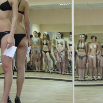 Still from Girl Model. Slightly-distorted mirror reflection of a line of six young girls who are dressed in bikinis and heels. They are standing against a wall of mirrors.