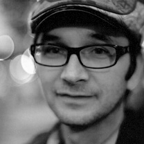 David Redmond looking straight ahead. He wears dark-rimmed glasses and a flat cap. Black and white portrait, with out of focus background.