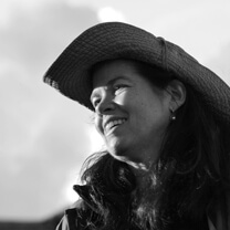 Pamela Yates looking to the side, smiling. She has long dark hair, and wears a wide-brimmed hat. Black and white portrait.