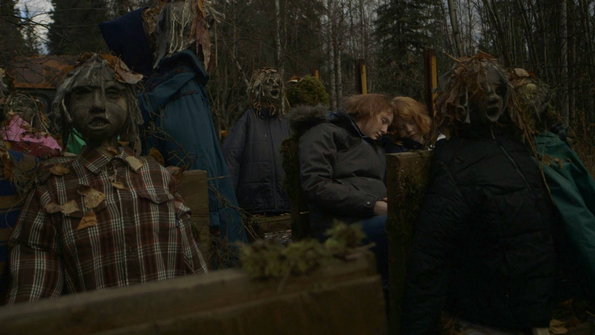 Still from LoveTrue. Two young folks are sitting and reclining their heads on each other, they wear winter jackets. They are surrounded by child-like manequins.