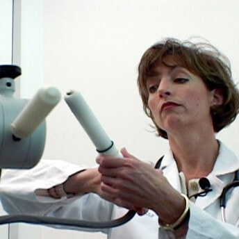 Still from Orgasm Inc. A woman in a medical jacket with a stethoscope around her neck and a mic lapel clipped to her jacket stands in front of a medical device. She is holding a piece of it.