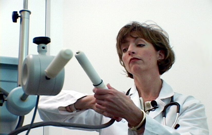 Still from Orgasm Inc. A woman in a medical jacket with a stethoscope around her neck and a mic lapel clipped to her jacket stands in front of a medical device. She is holding a piece of it.