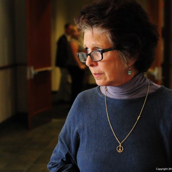 Still from Private Violence. A woman wearing glasses, a blue sweater, and a peace sign necklace is in a hallway looking away from the camera.