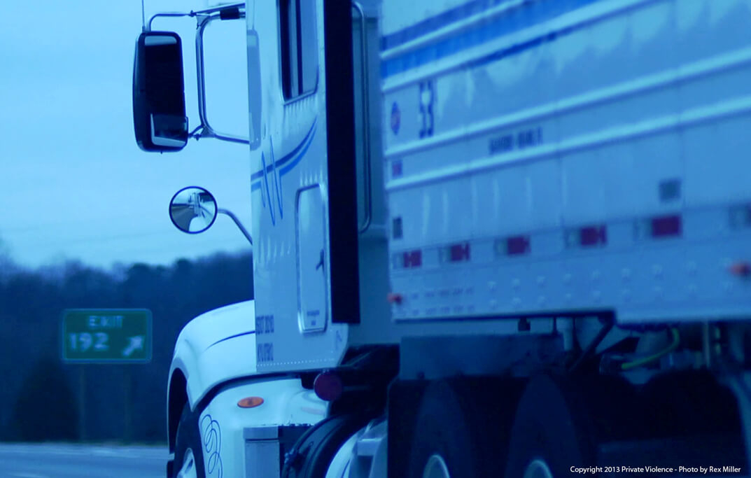 Still from Private Violence. Close-up of the side of a semi-tractor trailer truck. In the blurred background there is a green exit sign for "EXIT 192".