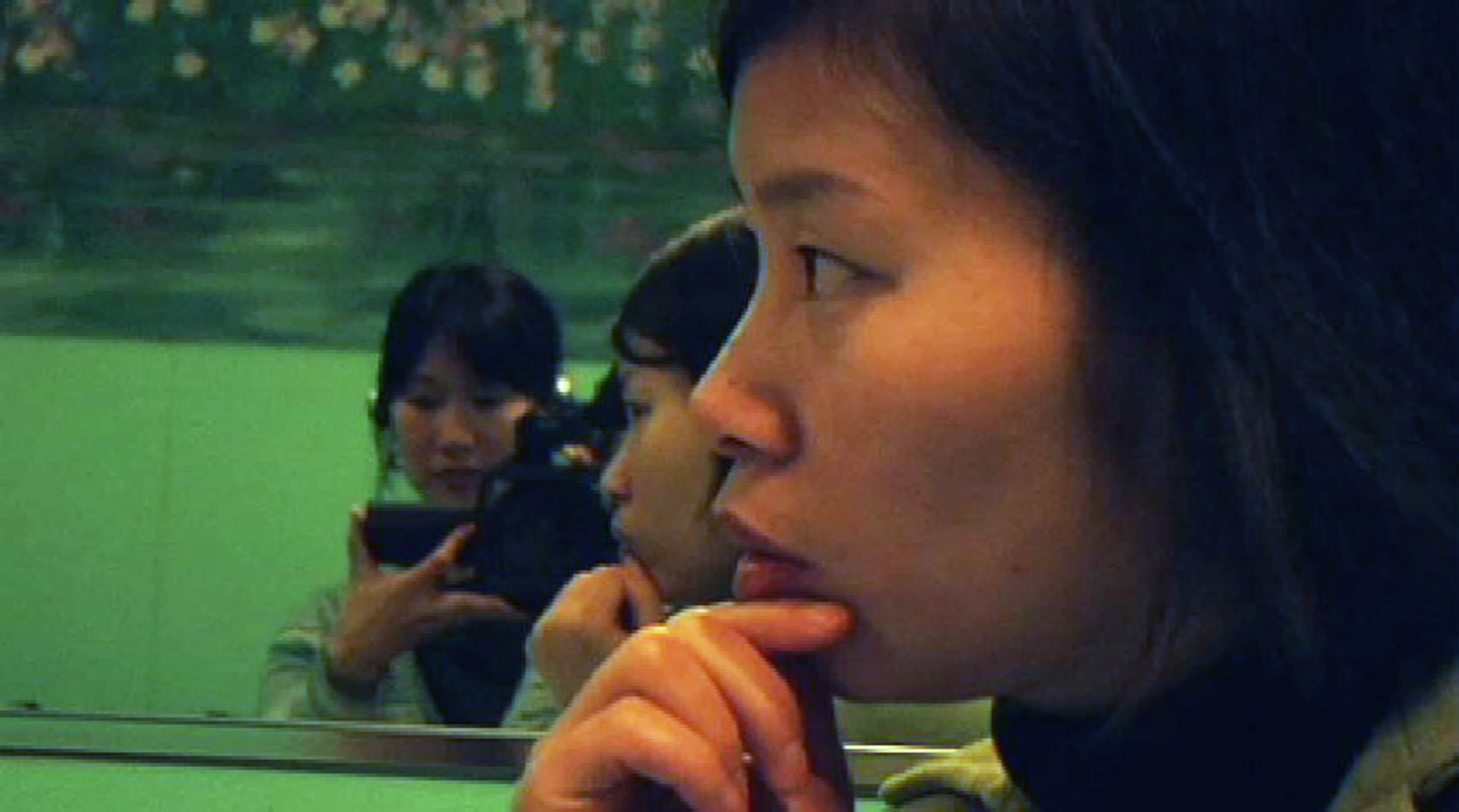 In the foreground a woman is looking away from the camera, her hand is touching her chin. In the background, there is a mirror reflection of Filmmaker Debbie Lum recording with a hand-held camera.