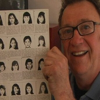 A smiling man holds a paper with both of his hands. The paper has the faces of Chinese women with descriptions below each photo. He wears eyeglasses and a headset.