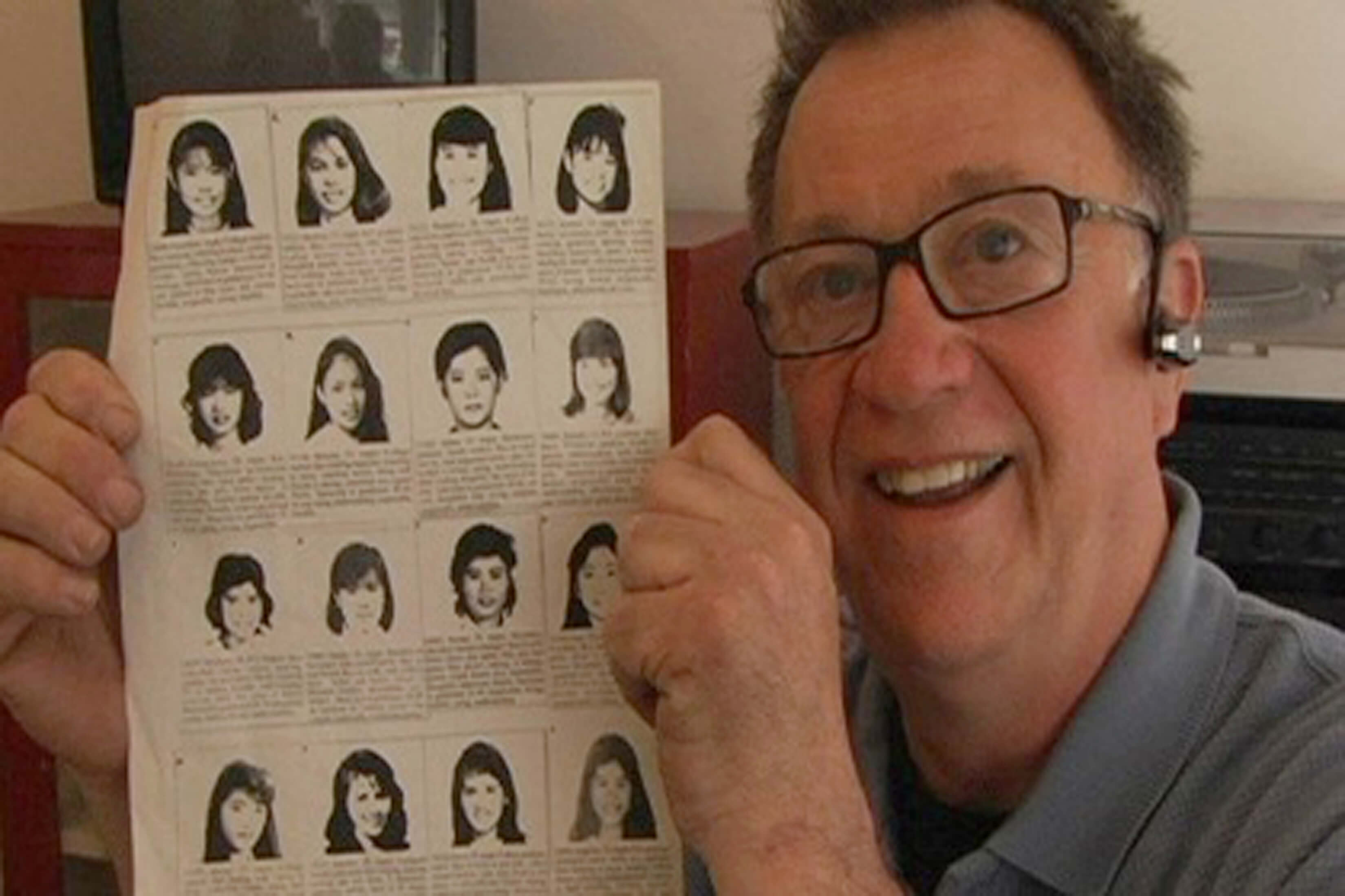 A smiling man holds a paper with both of his hands. The paper has the faces of Chinese women with descriptions below each photo. He wears eyeglasses and a headset.