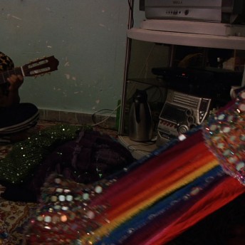 In the foreground, a woman with a colorful dress on top of her, in the background a person playing the guitar sitting on the floor.