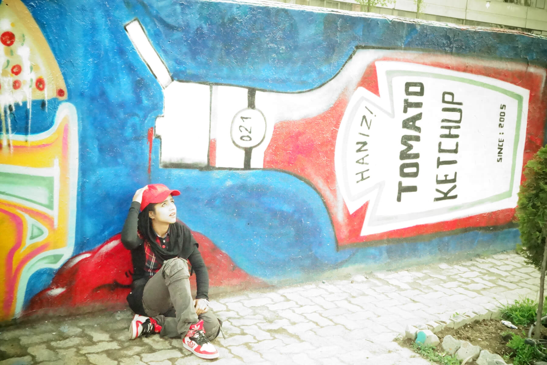 Sonita sitting on the ground in front of a wall with a mural of a ketchup bottle.